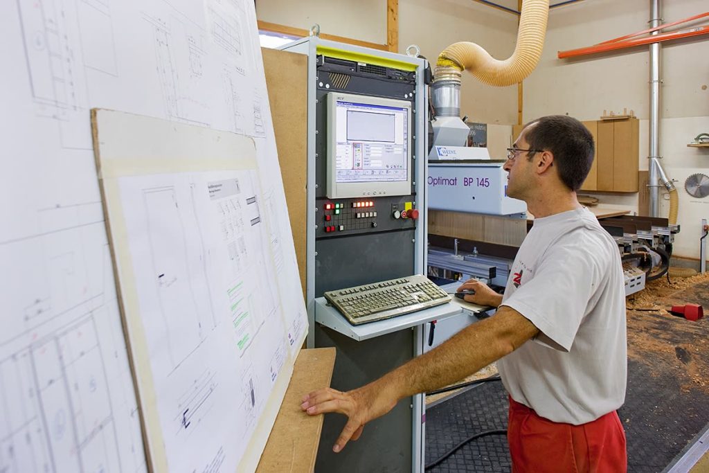 Mitarbeiter bedient die Steuerung einer CNC-Maschine neben einem aufgestellten Werkplan