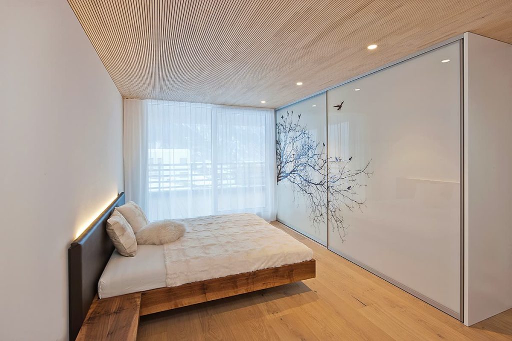 Schlafzimmer mit Holzbett, Lamellendecke und Schiebetüren aus Weissglas mit Baummotiv