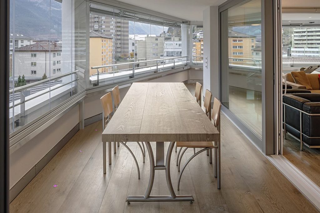 Langer Holztisch mit Stühlen auf einer verglasten Terrasse mit Blick auf die Stadt