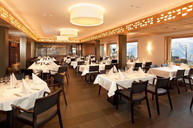 Restaurant mit gedeckten Tischen, dunklen Stühlen und Holzdecke mit durchbrochener Lichtblende – Zeiter + Berchtold, Brig-Glis