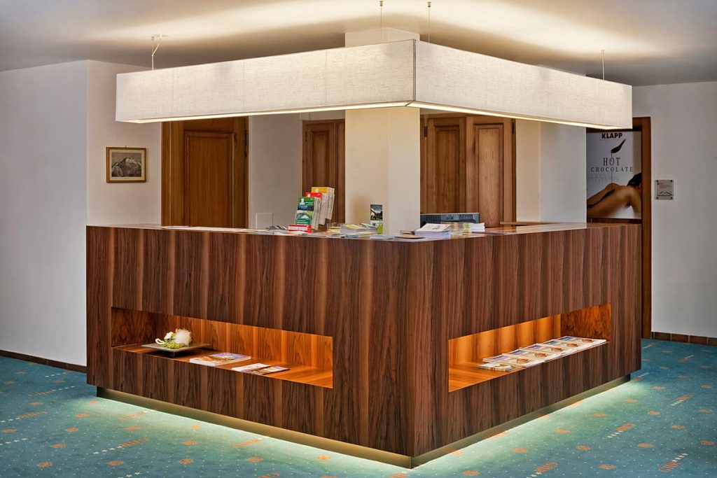 Empfangstheke aus Nussbaum mit beleuchteten Ablagefächern in einer Hotelhalle – Zeiter + Berchtold, Brig-Glis