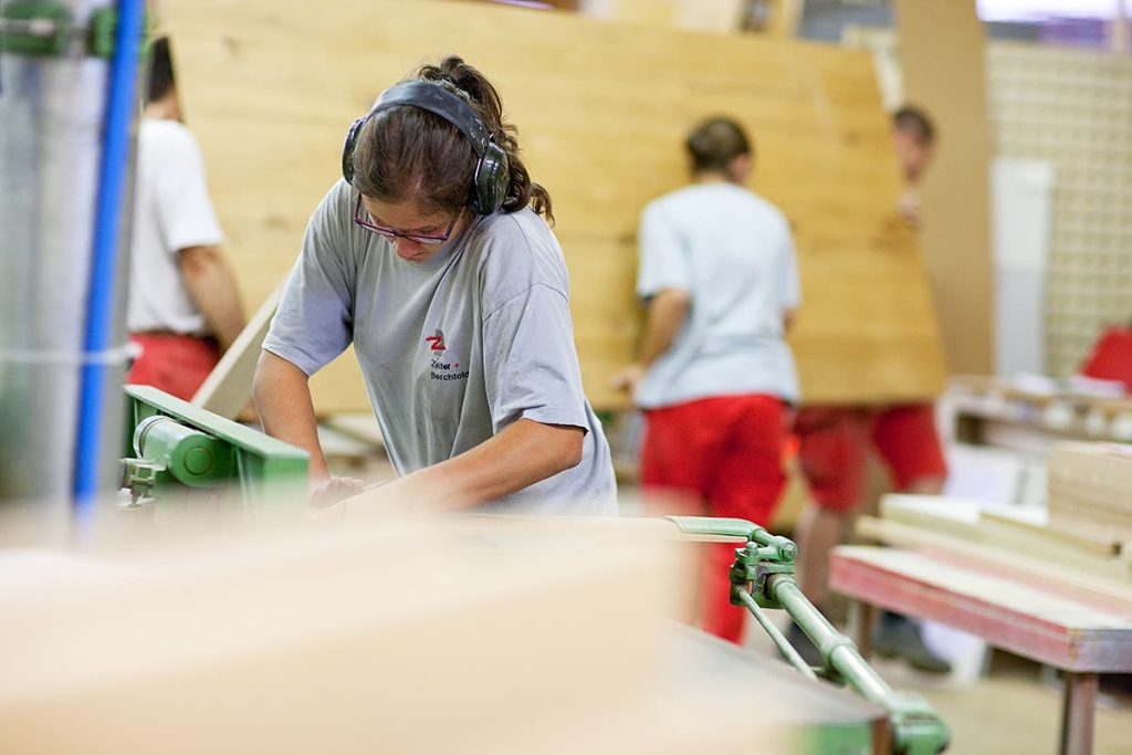 Mitarbeiterin mit Gehörschutz arbeitet in der Werkstatt, Kollegen tragen im Hintergrund eine Holzplatte