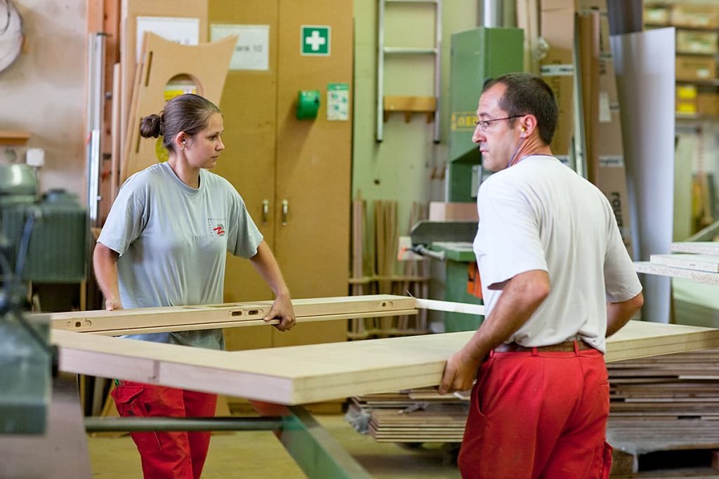 Zwei Mitarbeitende tragen eine lange Holzplatte durch die Werkstatt