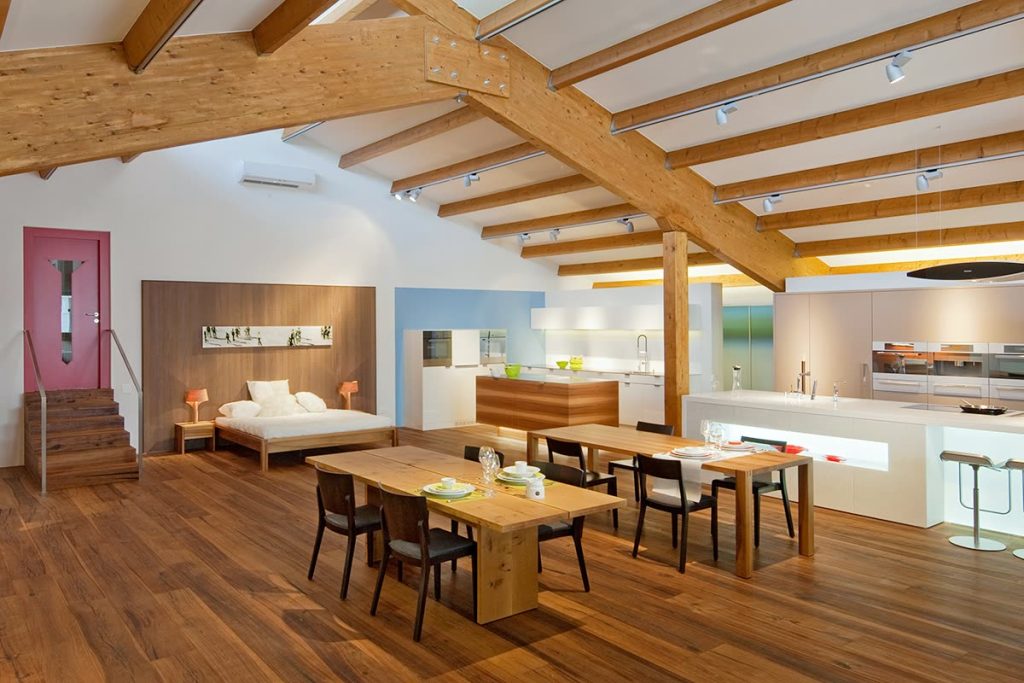 Showroom mit Holzbalken-Dachstuhl, Esstisch, Bett und Küche auf Parkett – Zeiter + Berchtold, Brig-Glis