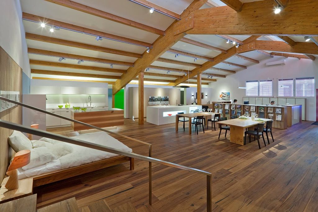 Weitläufiger Showroom unter sichtbarem Dachstuhl mit Bett, Esstisch und Küchen auf Parkettboden